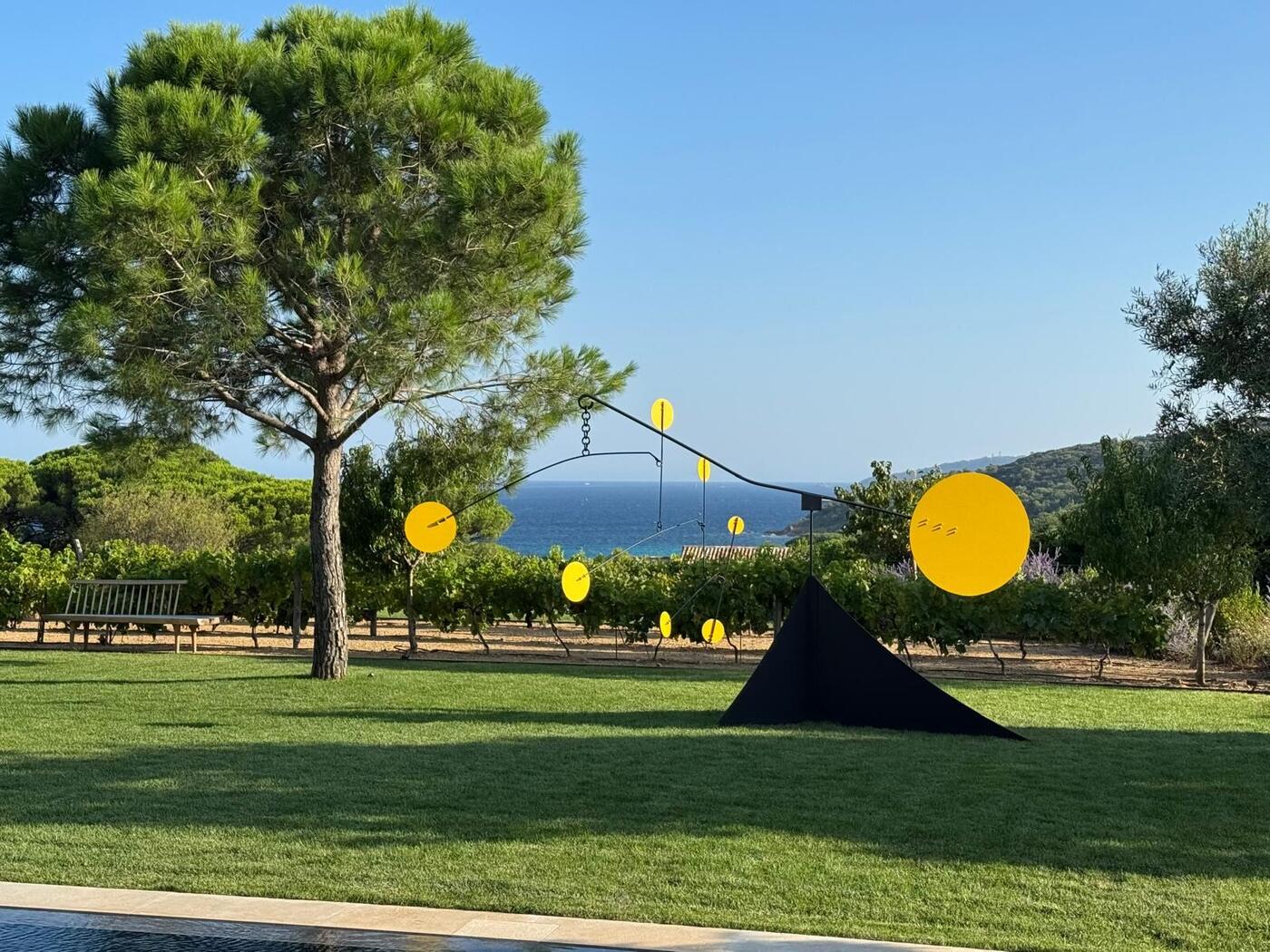 Le mobile Alfredo, pyramide noire et disques jaunes, sur une pelouse près d'un pin parasol avec la mer en fond